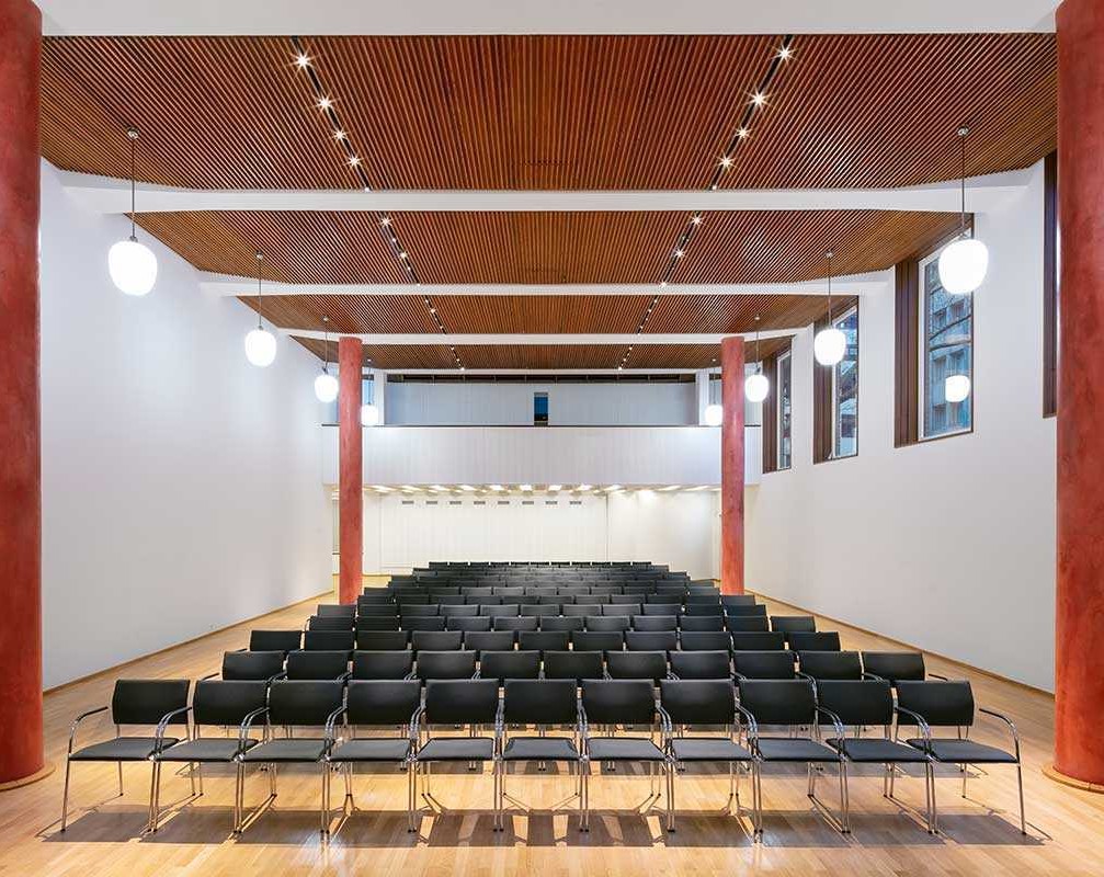 Ein Blick in den Overstolzensaal des MAKK: weiße Wände, eine Holzdecke, gesäumt von zwei roten Säulen auf jeder Seites des Raumes, eine Reihenbestuhlung mit schwarzen Stühlen.