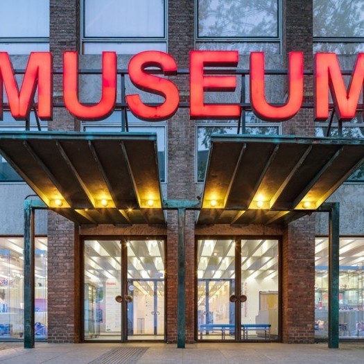 Ein frontales Foto des beleuchteten Eingangs des MAKK, über den Flügeltüren aus Glas ein beleuchtetes Vordach, darüber in großen roten Buchstaben das Wort "Museum".