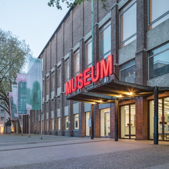 Das Gebäude des MAKK: ein roter Backsteinbau mit hohen Fenstern, im Zentrum der Eingang mit vorgelagertem Dach, darauf in großen roten Buchstaben das Wort "Museum".