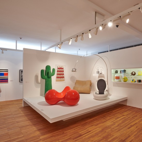 Ein Blick in die Ausstellung Kunst und Design im Dialog. Ein brauner Holzfußboden, weiße Ausstellungswände und ein weißes Podest, darauf verschiedene Designobjekte.