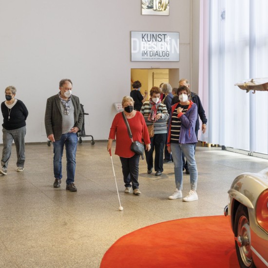 Eine Gruppe von Menschen mit Blindenstöcken läuft durch einen Ausstellungsraum des MAKK.