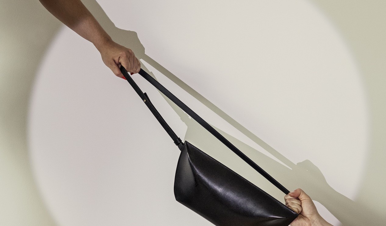 Schwarze Handtasche vor hellem Hintergrund. Zwei Hände ziehen an dem Schultergurt.