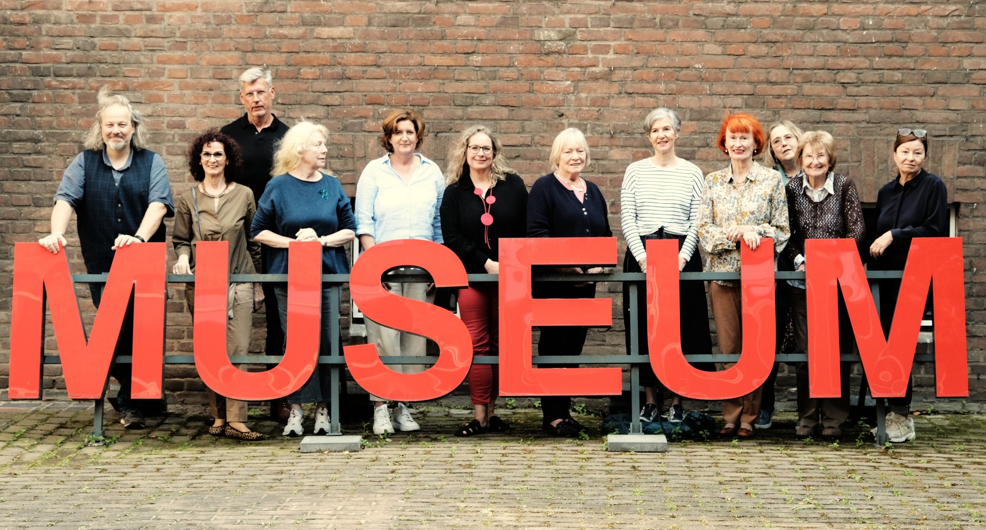 Der Arbeitskreis der Overstolzengesellschaft steht nebeneinander vor einer der Backstein-Außenwände des MAKK, vor ihnen rote Buchstaben: "Museum".