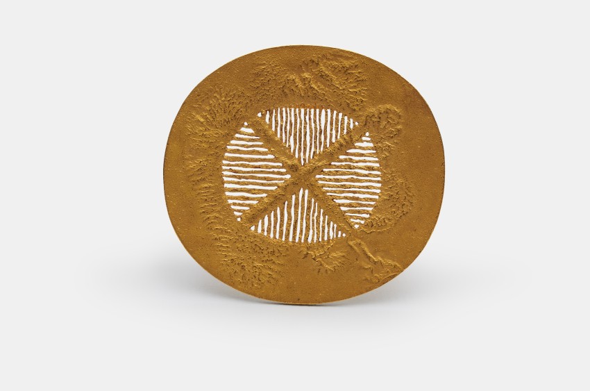 Leicht ovale, goldene Brosche. In der Mitte ein goldenes X mit goldener Gitterstruktur in den Zwischenräumen.