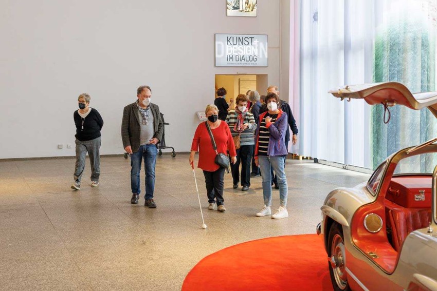 Eine Gruppe von Menschen mit und ohne Beeinträchtigungen besuchen einen Ausstellungsraum des MAKK, eine ältere Frau im Vordergrund führt einen Blindenstock vor sich.