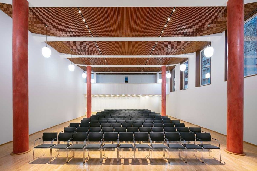 Ein Blick in den Overstolzensaal des MAKK: weiße Wände, eine Holzdecke, gesäumt von zwei roten Säulen auf jeder Seites des Raumes, eine Reihenbestuhlung mit schwarzen Stühlen.
