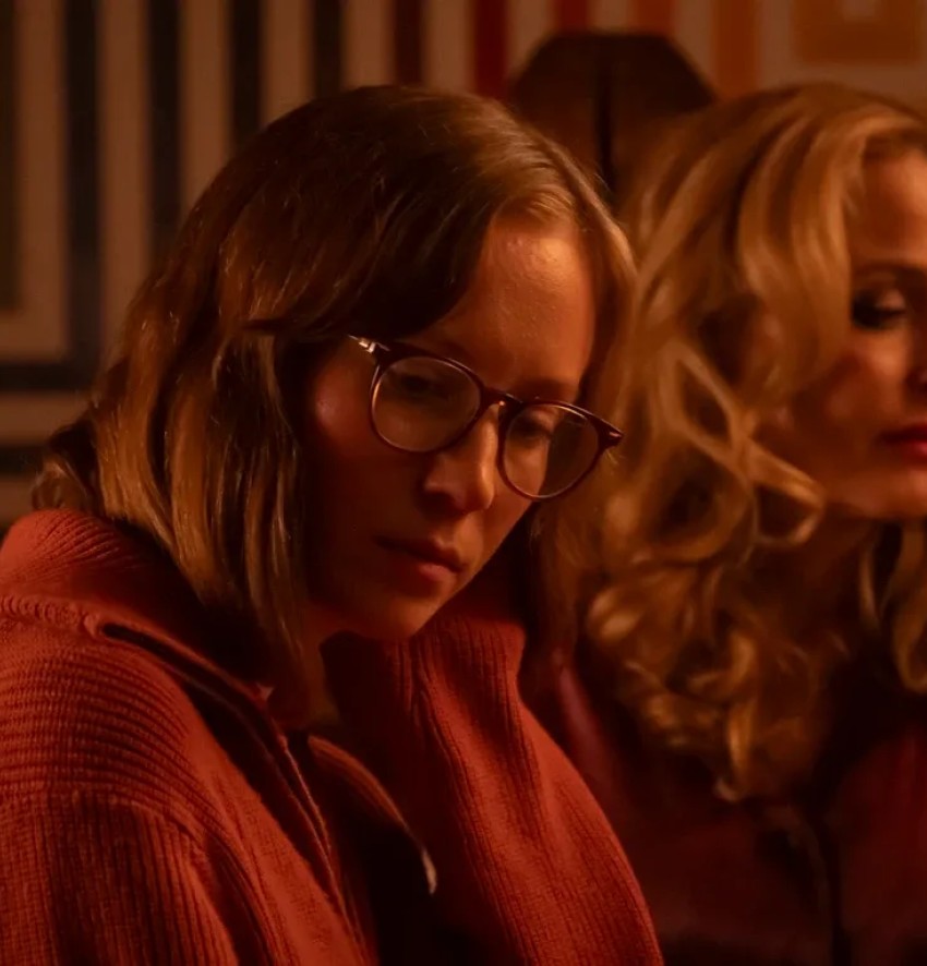 Eine atmosphärische, nahe Innenaufnahme aus dem Film „Teenage Sex And Death at Camp Miasma“. Im linken Vordergrund sitzt eine junge Frau mit Brille und mittellangen, braunen Haaren in einem dicken, rot-orangenen Strickpullover. Sie blickt mit gesenktem Kopf, nachdenklichem Ausdruck und leicht auf ihre Hand gestützt nach unten rechts. Direkt rechts hinter ihr sitzt eine ältere, blonde Frau mit elegant gewellten Haaren und rotem Lippenstift in einer glänzenden, rötlichen Seidenbluse, deren Blick unscharf in dieselbe Richtung geht. Der Hintergrund ist von einer Wand mit einem geometrischen, quadratischen Muster in warmen Erd- und Rottönen sowie einer hölzernen Treppenstruktur geprägt. Die gesamte Szene ist in ein sehr warmes, gedimmtes, rot-oranges Licht getaucht, das eine vertraute, aber auch leicht melancholische oder angespannte Stimmung erzeugt.