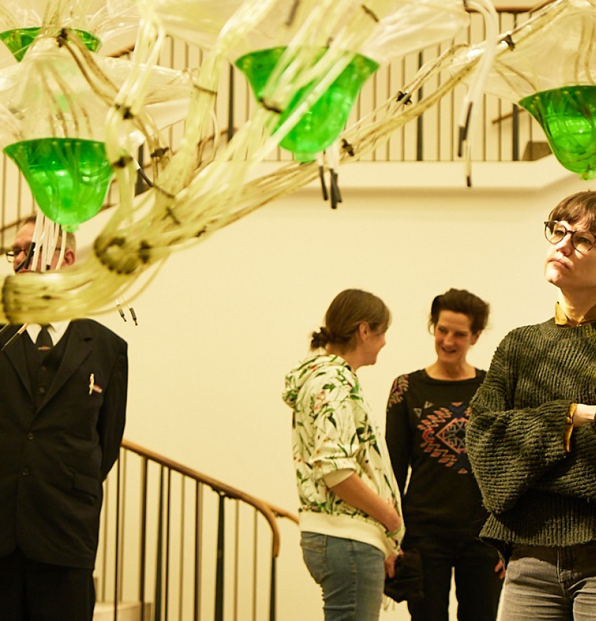 An installation with green glass elements suspended from a scaffold, behind it a young woman looking at the object. Behind her a small group of people are deep in conversation.