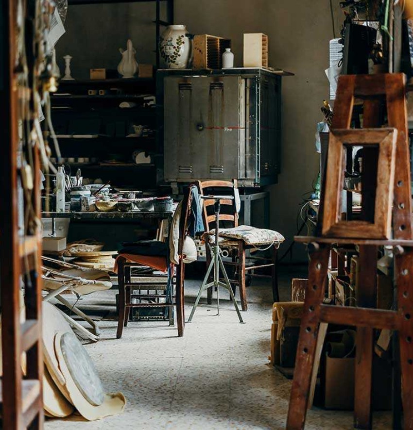 A very messy workshop, on the left shelves with various objects, in the background a large metal tub, in front of it some chairs.