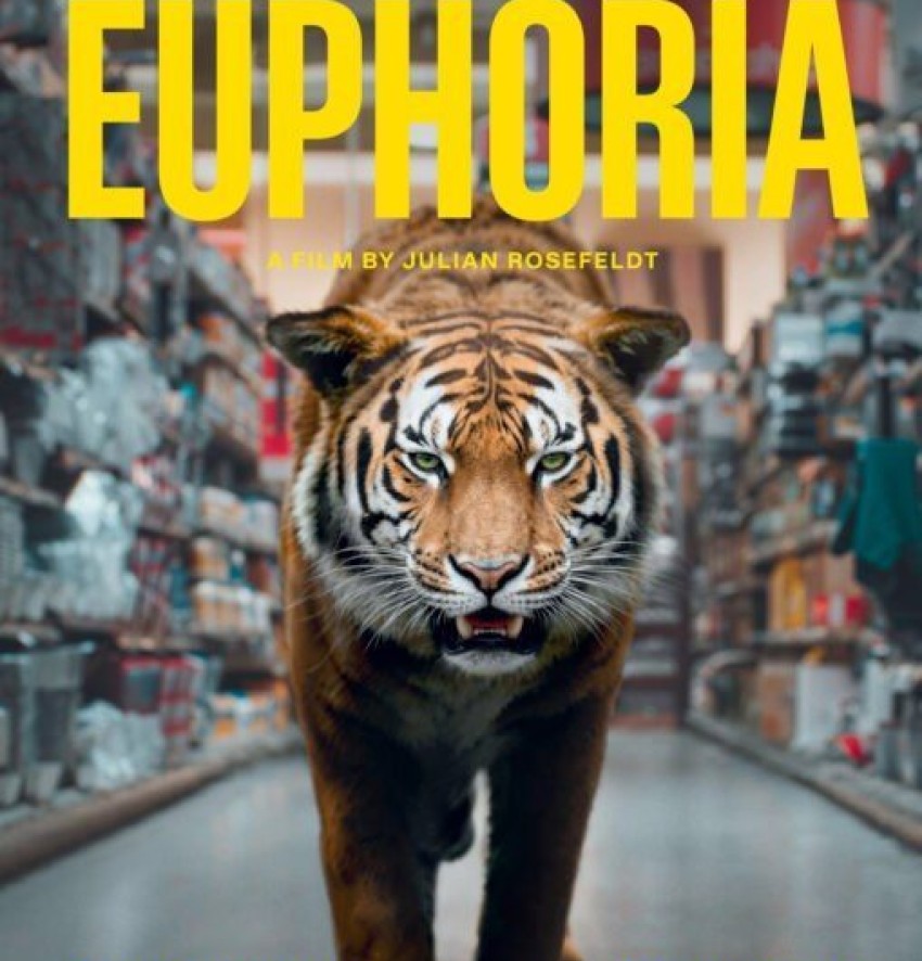 Eine frontale, zentrierte Aufnahme eines majestätischen Tigers, der direkt auf den Betrachter zugeht. Das Raubtier befindet sich in einem endlos wirkenden, langen Supermarktgang, der links und rechts von hohen, vollgepackten Regalen gesäumt ist. Der Tiger fixiert den Betrachter mit einem intensiven, grünen Blick aus leicht zusammengekniffenen Augen, sein Maul ist drohend geöffnet und zeigt die Zähne. Das detaillierte, orange-schwarze Fell des Tieres steht im starken Kontrast zur künstlichen, sterilen Umgebung des Kaufhauses. Die Gänge und die Deckenbeleuchtung erzeugen eine tiefe Perspektive, die das Tier dramatisch in Szene setzt. Am oberen Bildrand dominiert in großen, leuchtend gelben Großbuchstaben der Filmtitel „EUPHORIA“. Darunter steht in kleinerer, gelber Schrift „A FILM BY JULIAN ROSEFELDT“. Im unteren Drittel, auf Höhe der Beine des Tigers, sind die Namen der Hauptdarsteller in gelber Schrift platziert: links „GIANCARLO ESPOSITO“ und rechts „CATE BLANCHETT“. Ganz am unteren Rand befindet sich der übliche, detaillierte Credit-Block in sehr kleiner Schrift. Die surrealistische Bildkomposition fängt einen Moment der ungezähmten Wildheit inmitten der modernen Konsumgesellschaft ein.