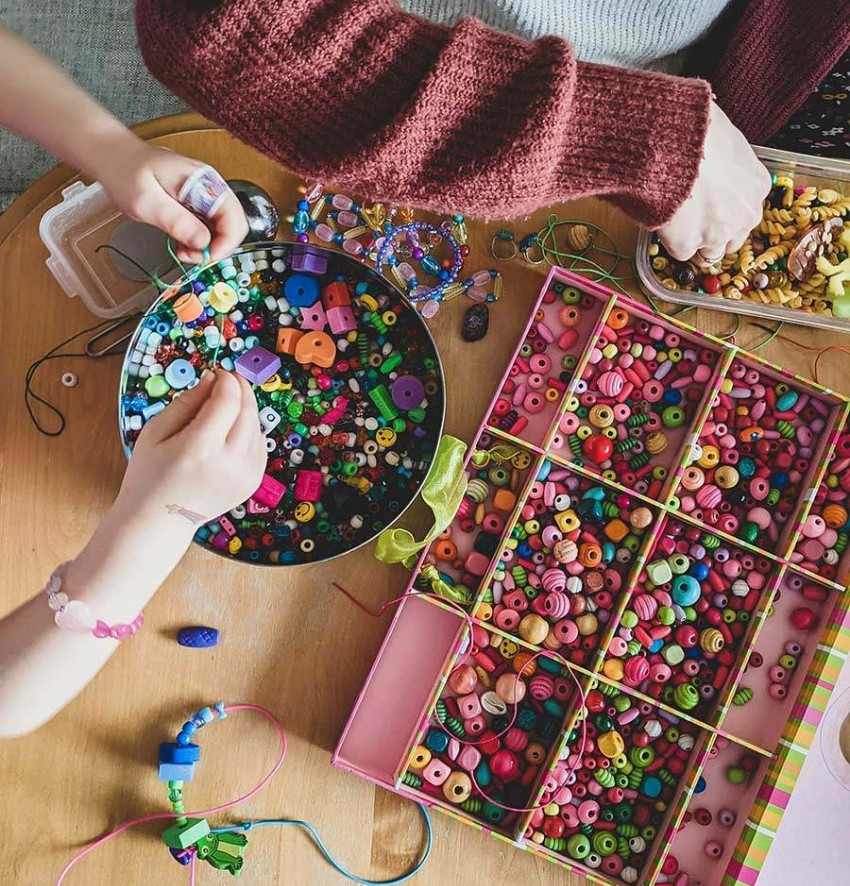 Ein Blick von oben: Zwei Kinder an einem Tisch, die aus verschiedenen Perlen und ähnlichen Objekten Schmuck basteln.