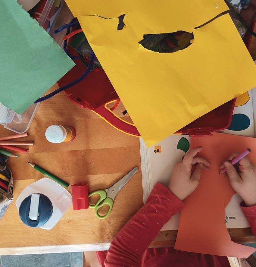 Ein Blick von oben auf einen Tisch mit vielen Bastelmaterialien. Zwei Kinderhände, die auf einem Stück Tonpapier malen.