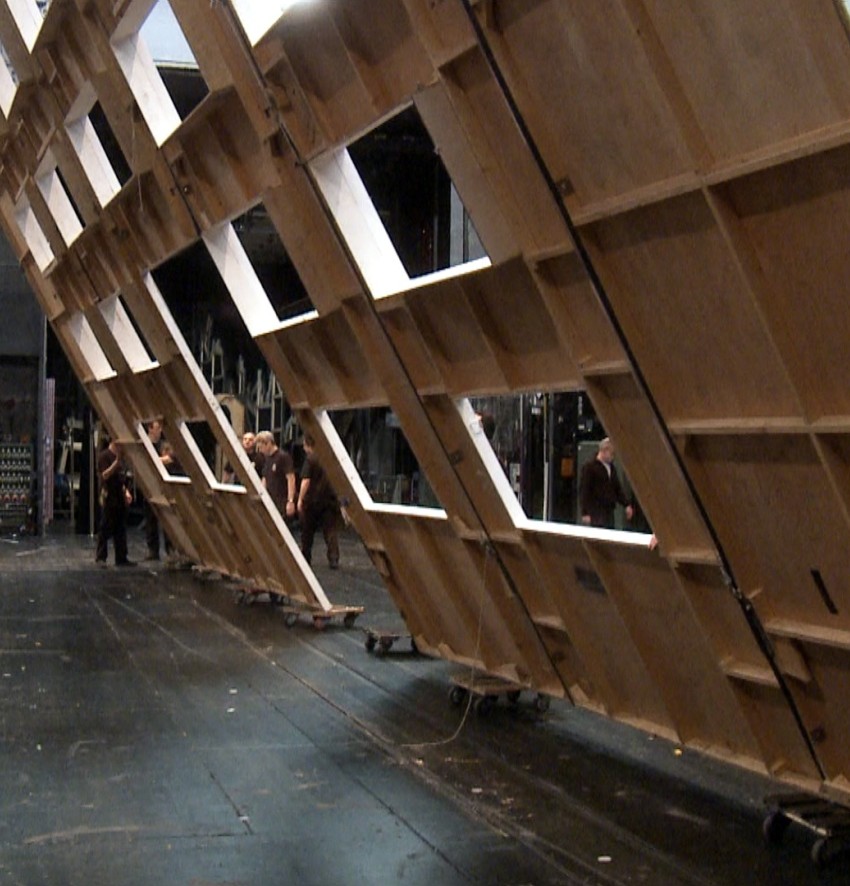 Eine fotografische Einblickaufnahme aus dem Blickwinkel der Hinterbühne eines großen Opernhauses während des Kulissenaufbaus. Ein massives, rahmenartiges Holzfenster-Bühnenelement mit rechteckigen Aussparungen wird schräg von mehreren Bühnenarbeitern nach oben gehoben oder gekippt. Im linken Vordergrund geht ein Techniker in dunkler Kleidung über den glänzenden, dunklen Bühnenboden, während rechts im Vordergrund ein weiterer Arbeiter in Latzhose von hinten zu sehen ist. Der Hintergrund gibt den Blick frei auf die komplexe, mehrstöckige Bühnentechnik und Werkstattstruktur des Hauses mit dunklen Metallgerüsten, Scheinwerferbrücken, Zügen und vollgepackten Regalsystemen an den Seitenwänden. Die Atmosphäre wirkt geschäftig, technisch und gewaltig.