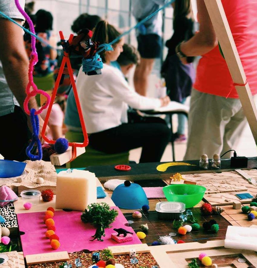 A creative workshop setting: In the foreground a table with various colorful craft materials, behind it a group of people.