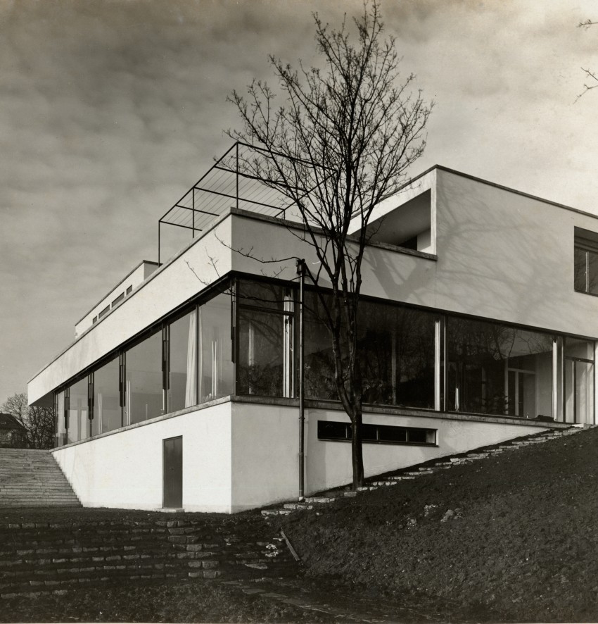 Eine Schwarz-Weiß-Fotografie einer modernen Villa im Stil der klassischen Moderne auf einem leicht abfallenden Grundstück. Das mehrstöckige, kubische Gebäude besticht durch seine schneeweißen, glatten Fassaden und eine riesige, durchgehende Glasfront im Erdgeschoss, die den Blick ins lichtdurchflutete Innere freigibt. Links führt eine breite Steintreppe zu einer schattigen Terrasse hinauf, während rechts ein begrünter Hang an das Haus grenzt. Ein einzelner, laubloser Baum steht direkt vor der verglasten Front und wirft filigrane Schatten auf die weiße Hauswand. Auf dem flachen Dach zeichnet sich eine quaderförmige Metallstruktur aus dünnen Streben ab. Der Himmel im Hintergrund ist leicht bewölkt und verleiht der minimalistischen Architektur eine ruhige, zeitlose Atmosphäre.