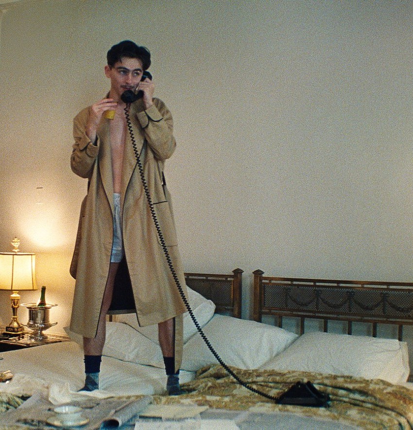 Ein dynamisches, leicht skurriles Szenenbild aus einem eleganten Hotelzimmer des A24-Films „Marty Supreme“. Im linken Bildzentrum steht der Schauspieler Timothée Chalamet barfuß auf einem großen Doppelbett. Er trägt einen langen, beigefarbenen Trenchcoat oder Bademantel, der vorne offen ist und den Blick auf seinen nackten Oberkörper und eine hellblaue Boxershorts freigibt. Er hält den Hörer eines schwarzen Oldtimer-Telefons mit langem, spiralförmig geschwungenem Kabel an sein Ohr und telefoniert mit konzentriertem Blick. Das Bett hinter ihm hat ein klassisches, hölzernes Kopfteil und ist mit weißen Kissen ausgestattet; im Vordergrund ist das Bett unordentlich mit Decken, Zeitungen und einem Frühstückstablett bedeckt. Links und rechts neben dem Bett steht jeweils ein Nachttisch mit einer leuchtenden, klassisch verzierten Tischlampe. Im linken Hintergrund ist eine prachtvolle, weiße Kassettentür einen Spalt breit geöffnet. Das Bild hat einen körnigen, warmen Retro-Look, der an die Mitte des 20. Jahrhunderts erinnert.