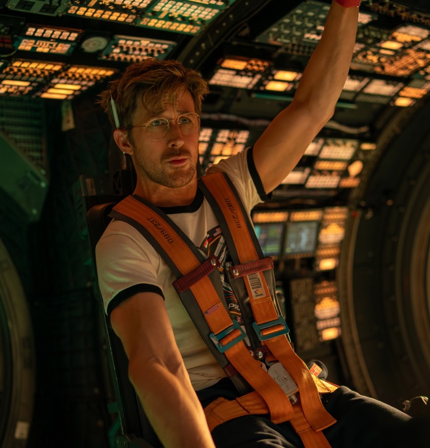 Ein actionreiches, halbnahes Szenenbild aus dem Science-Fiction-Film „Project Hail Mary“. Im Zentrum sitzt der Schauspieler Ryan Gosling als Astronaut Ryland Grace in einem Pilotensitz im Cockpit eines Raumschiffs. Er trägt ein weißes T-Shirt mit schwarzen Bündchen, eine feine Brille und hat einen Stift hinter sein Ohr geklemmt. Er ist mit breiten, leuchtend orangefarbenen Sicherheitsgurten fest im Sitz angeschnallt. Sein linker Arm ist nach oben gestreckt, um ein Bedienelement an der Decke zu erreichen, während sein Blick konzentriert und leicht besorgt nach vorne gerichtet ist. Das Cockpit um ihn herum ist kreisrund geformt, extrem detailreich und vollgepackt mit unzähligen leuchtenden Knöpfen, Kontrollpaneelen und Bildschirmen. Unten rechts ist ein kleiner Monitor mit grünen Grafiken zu sehen. Die Szene wird von einem warmen, gedimmten orange-grünen Licht dominiert, das eine angespannte und technologische Atmosphäre erzeugt.