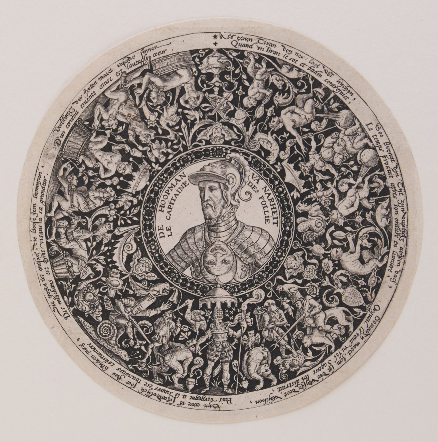 Kreisförmige Stichvorlage für den Boden einer Schüssel. In rundem Rahmen ist mittig das Portrait eines Mannes zu sehen, spiegelverkehrt unter ihm eine Narrenmaske. Rings um ihn herum sind auf schwarzem Grund zahlreiche Figuren, Tiere, Pflanzen und Zierelemente abgebildet.