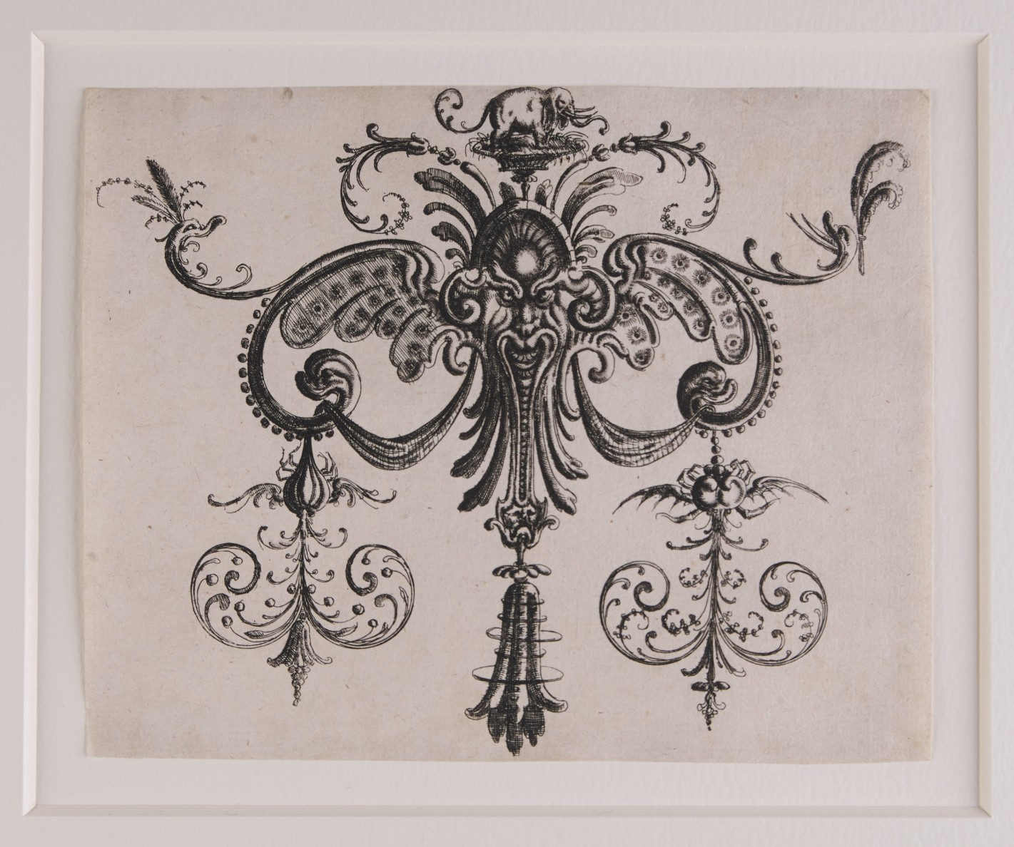 Verschlungene Ornamentform, mittig eine Fratze über der ein kleiner Elefant thront.